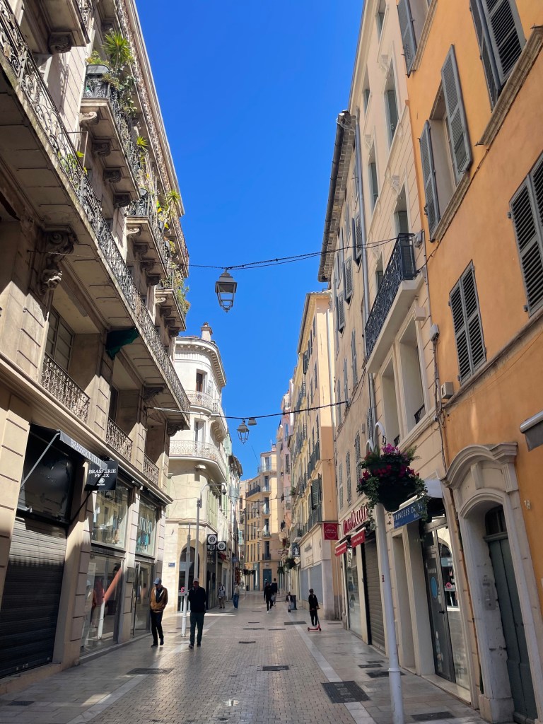 Úzká ulice v Toulonu s historickými budovami, modrou oblohou a několika chodci.