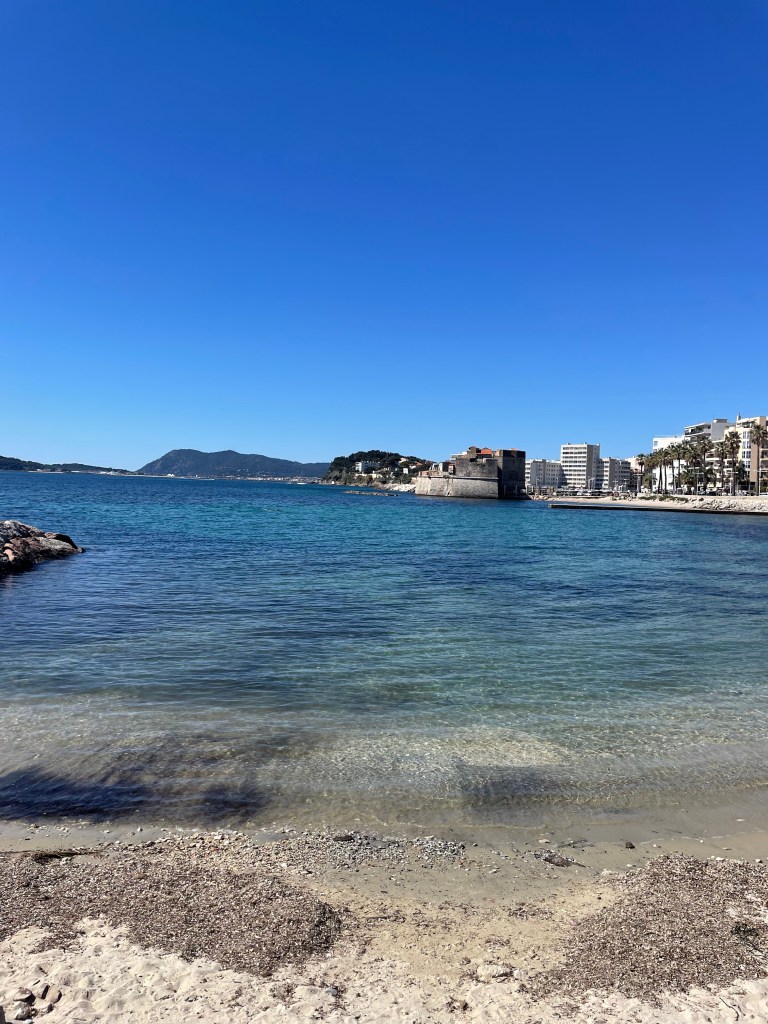 Pohled na pobřeží města Toulon s písečnou pláží a modrou vodou pod jasnou oblohou.