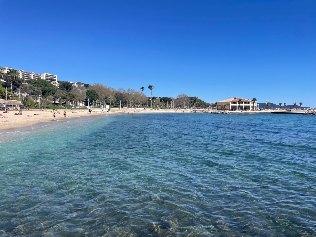 Pohled na písečnou pláž v Toulonu s modrou vodou a palmami, v pozadí budovy a slunečné nebe.