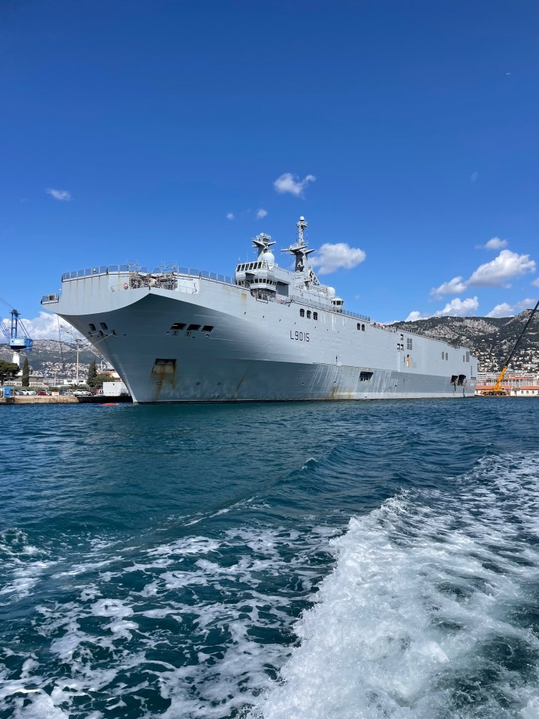 Loď ve vojenském přístavu v Toulonu, s modrou oblohou a mořem v pozadí.